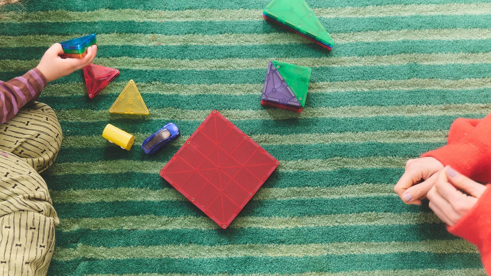 A child holding colorful magnetic tiles beside an adult on a striped rug.