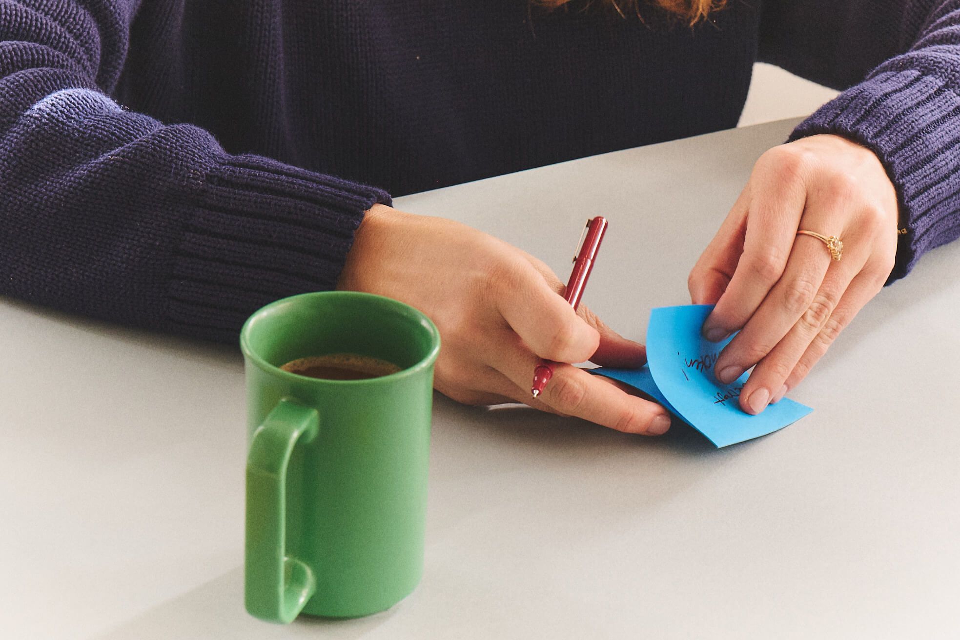 Hands writing on a blue sticky note at a desk, next to a coffee mug