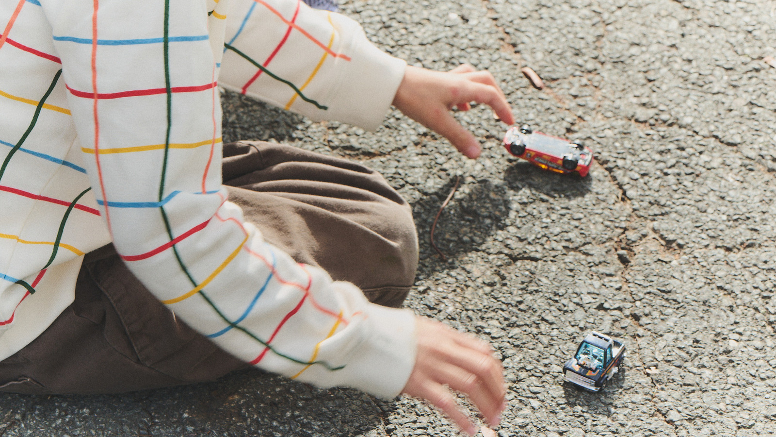 A child in a striped sweatshirt plays with two toy cars on pavement.