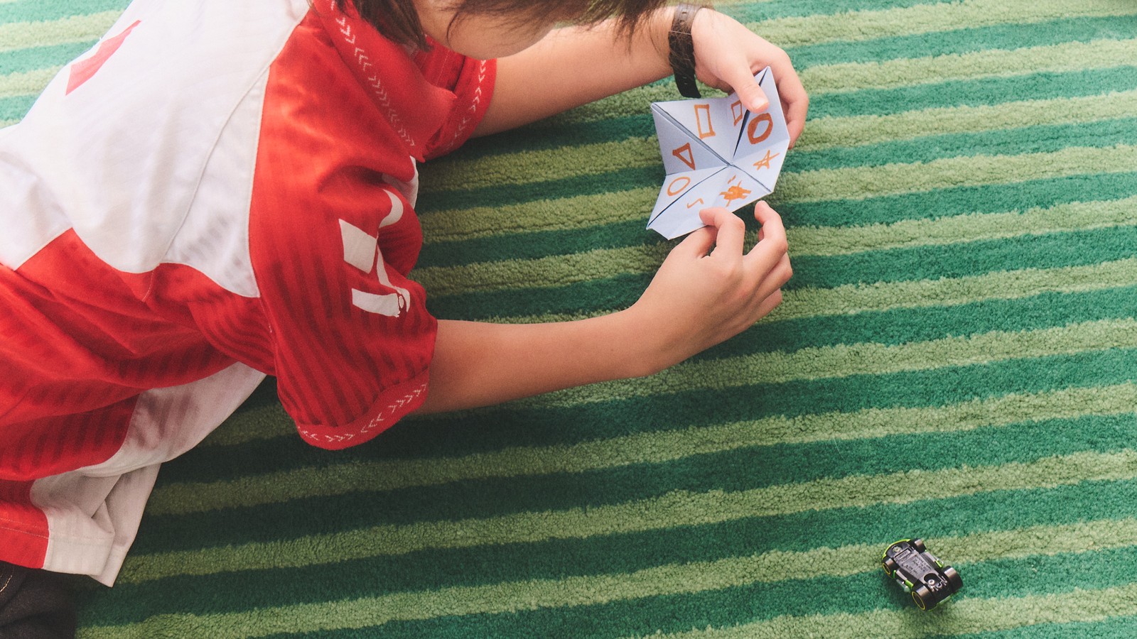 A child holds a folded paper fortune teller while wearing a watch on a green striped rug.
