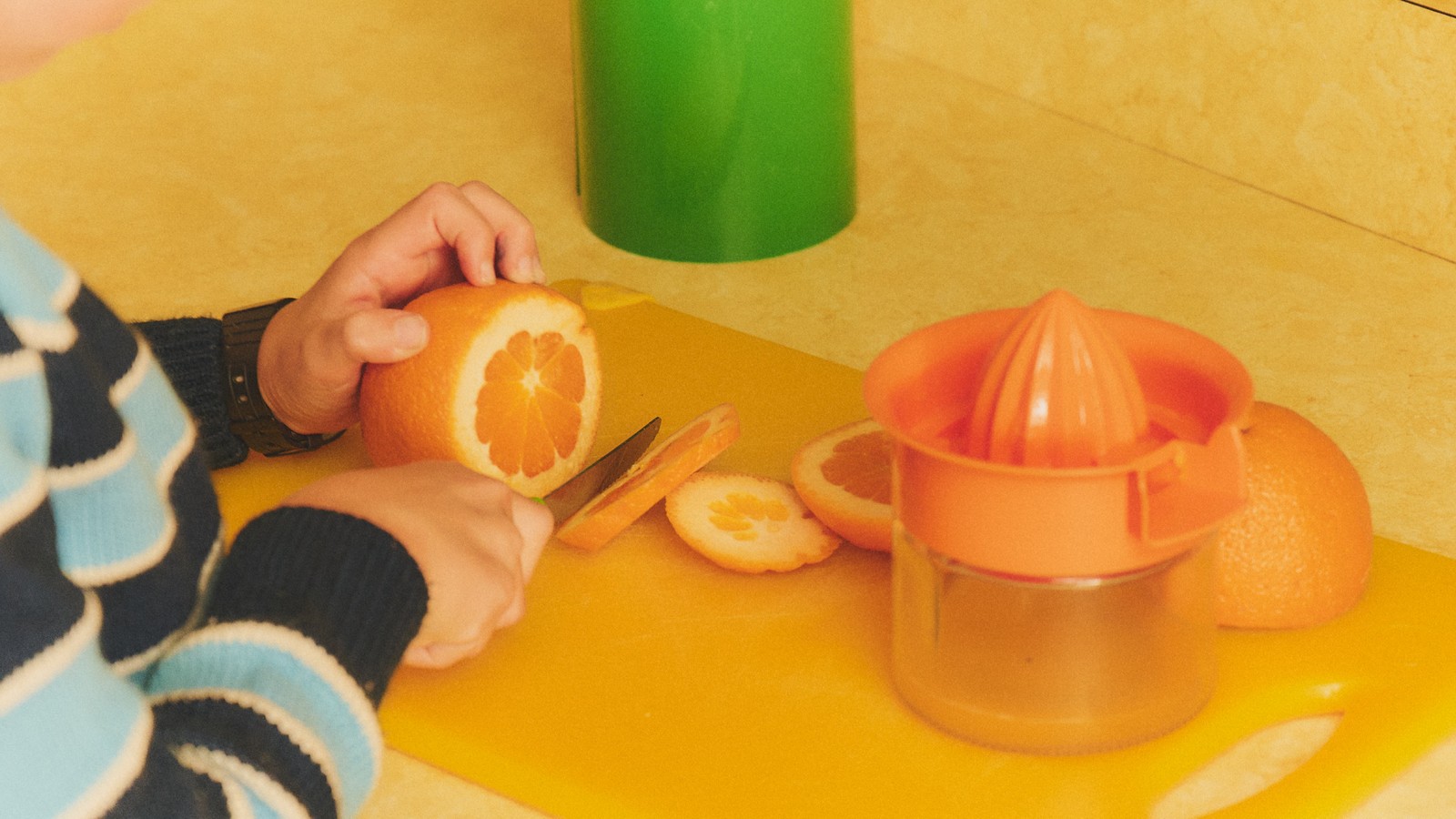 A child's hands slice an orange beside a citrus juicer on a yellow counter.