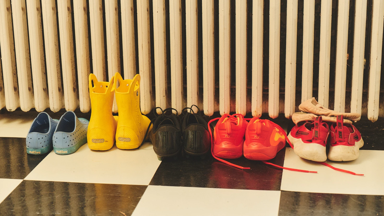 Pairs of clogs, rain boots, sneakers, and ballet slippers line up on a checkered tile floor.
