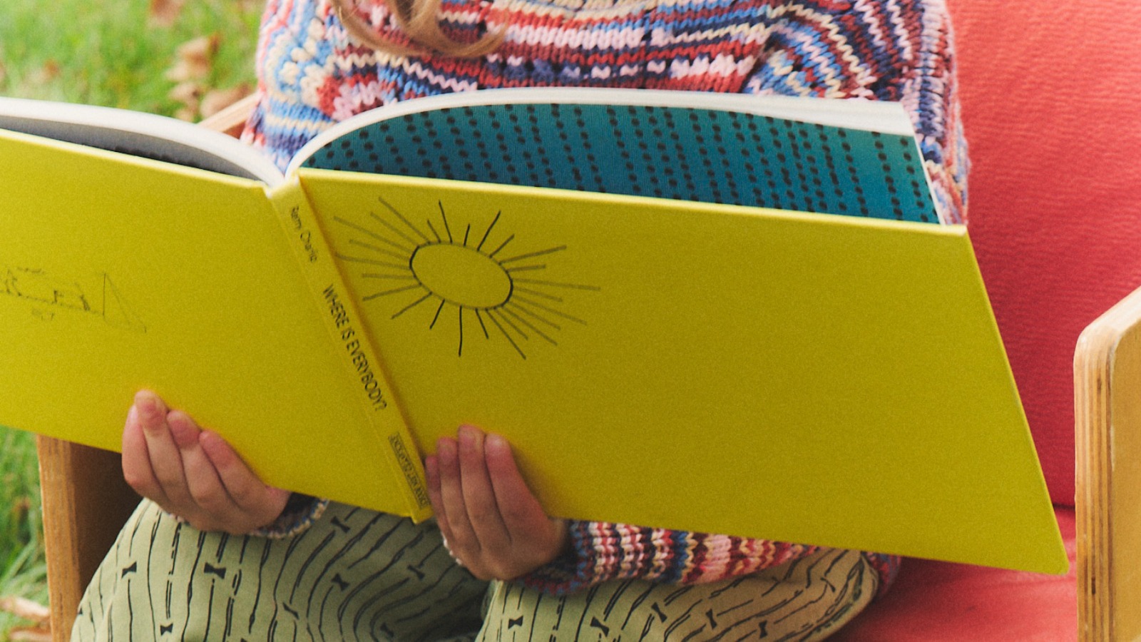 A child seated outdoors holds open a large yellow picture book.